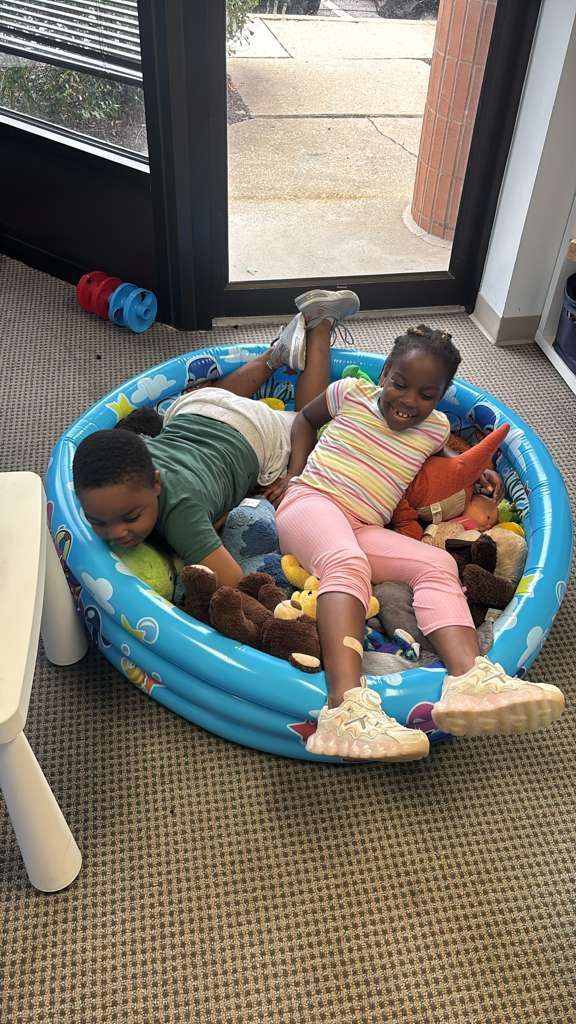 Two children in a blue inflatable pool filled with stuffed animals. 