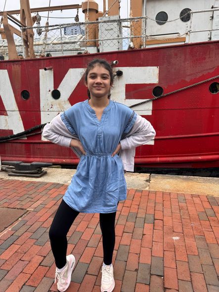 Girl posing in front of a red ship, wearing denim dress, black leggings, and white sneakers.