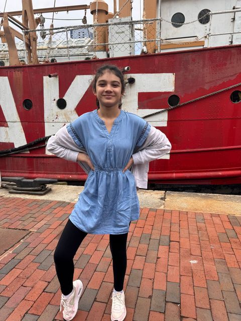 Girl posing in front of a red ship, wearing denim dress, black leggings, and white sneakers.