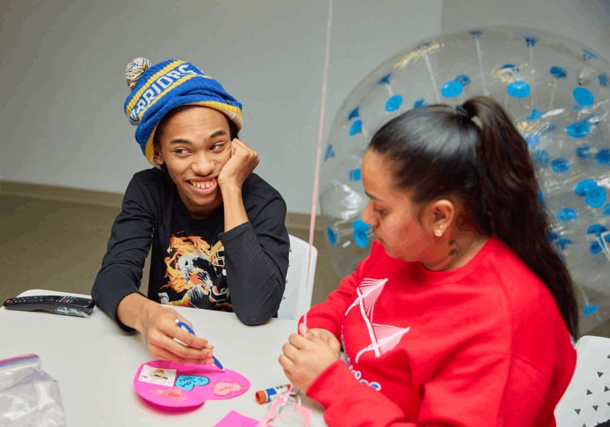 Person wearing hat smiles at another person at a table, both interacting with items; room setting.