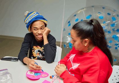 Person wearing hat smiles at another person at a table, both interacting with items; room setting.