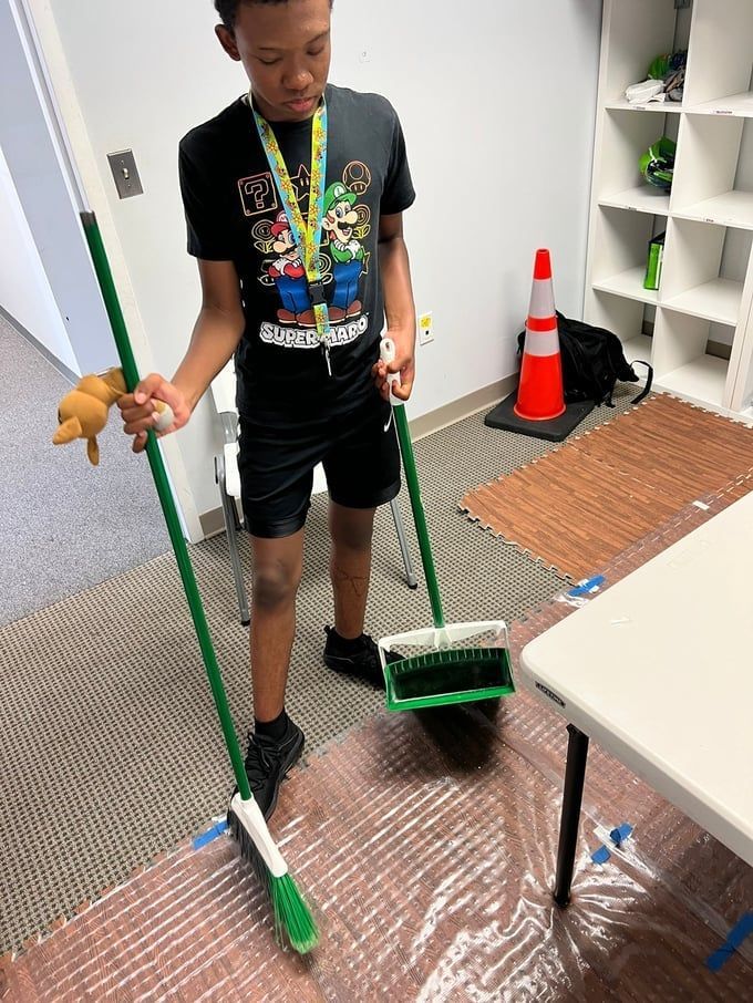 Person sweeps with two brooms in an office, wearing a Mario t-shirt, shorts, and sneakers.