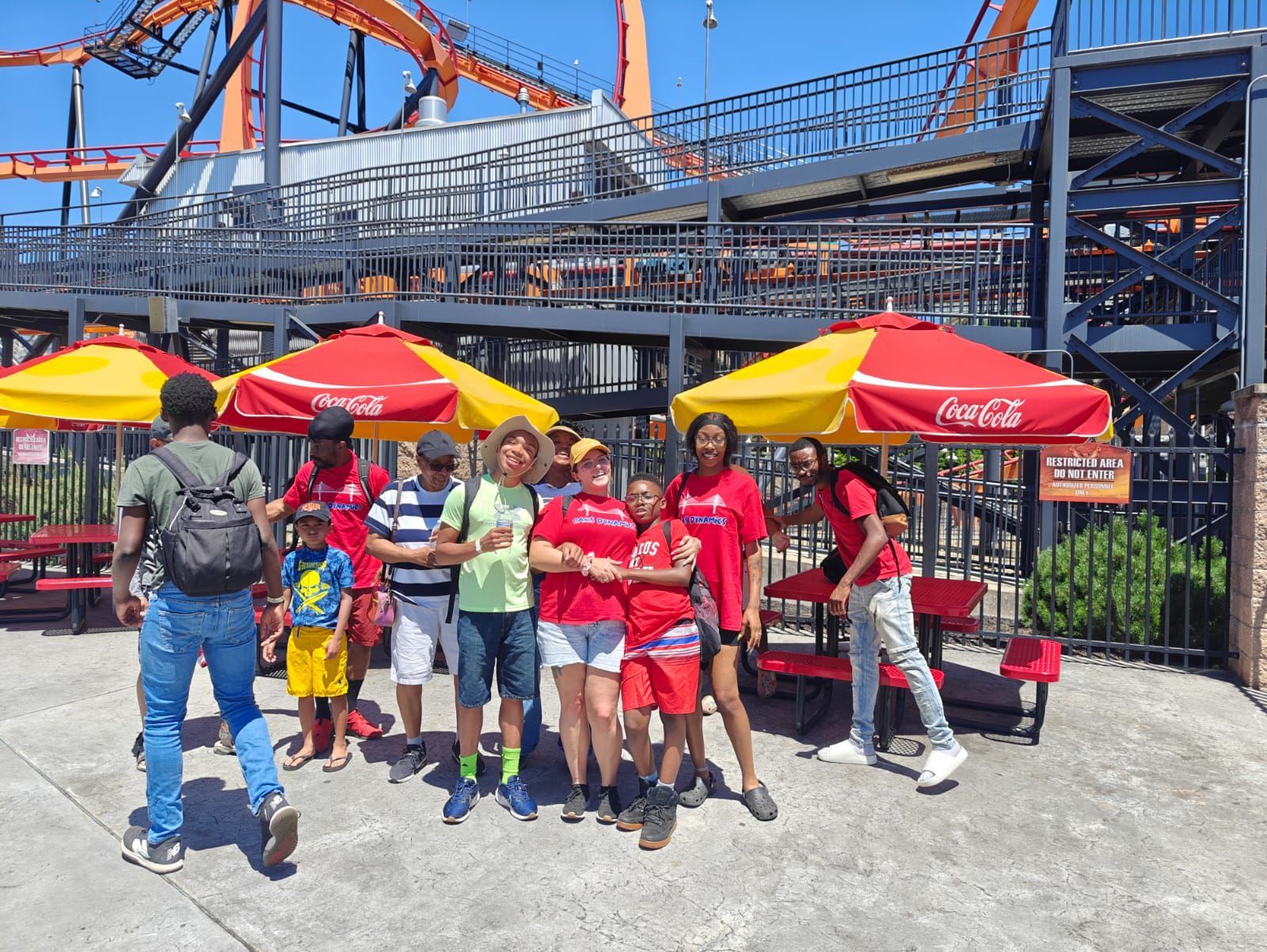 Group of people pose near a roller coaster with Coca-Cola umbrellas.