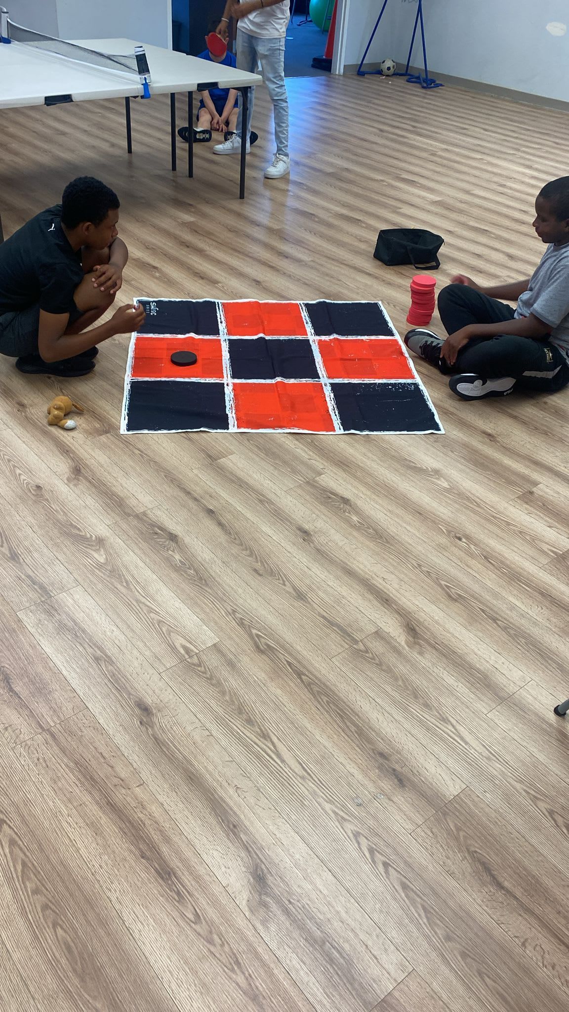 Two people playing checkers on a large board on a wooden floor indoors.