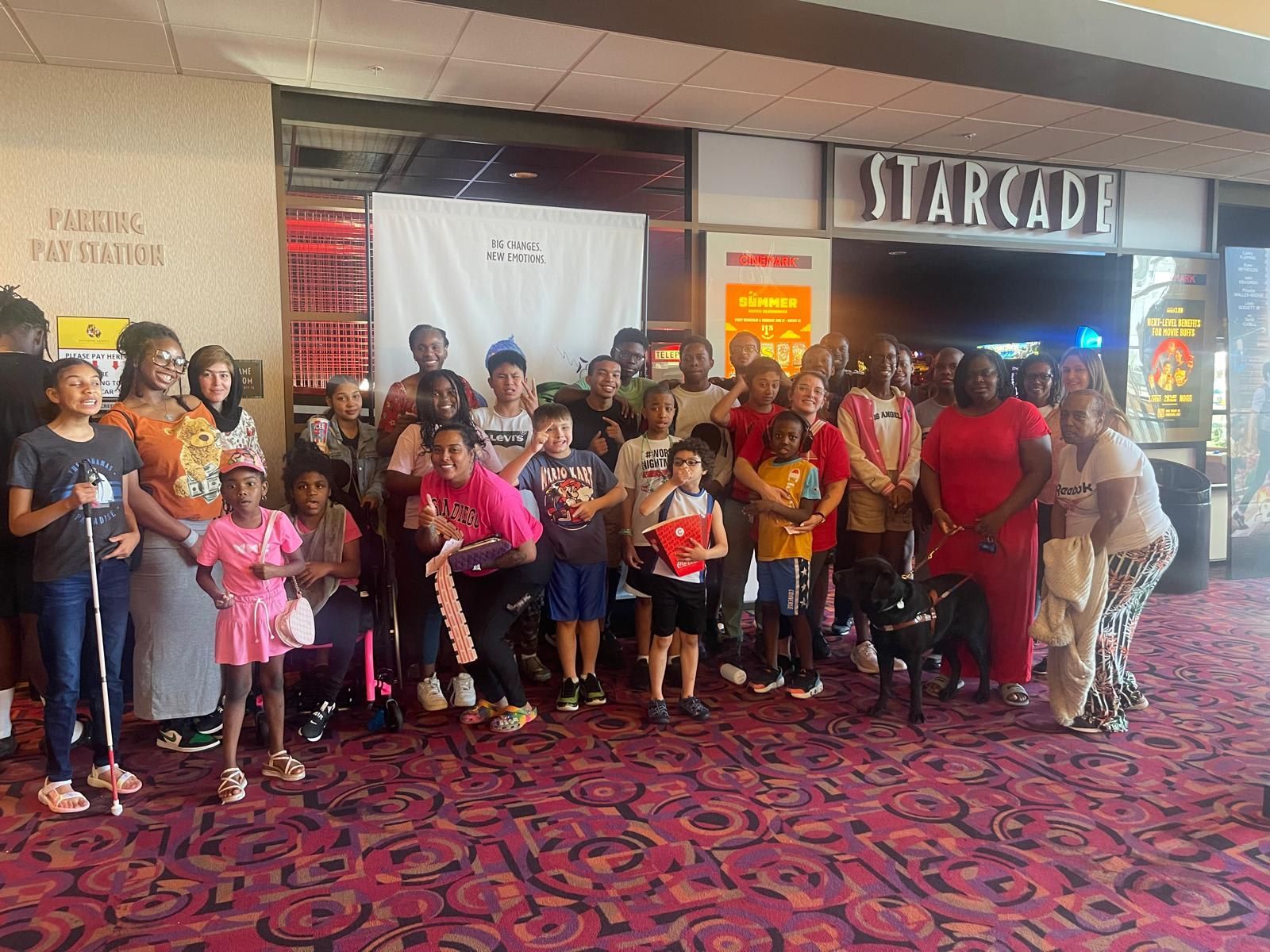 Group of people in front of a movie theater with a “STARCADE” sign. Many children are present. Red carpet.