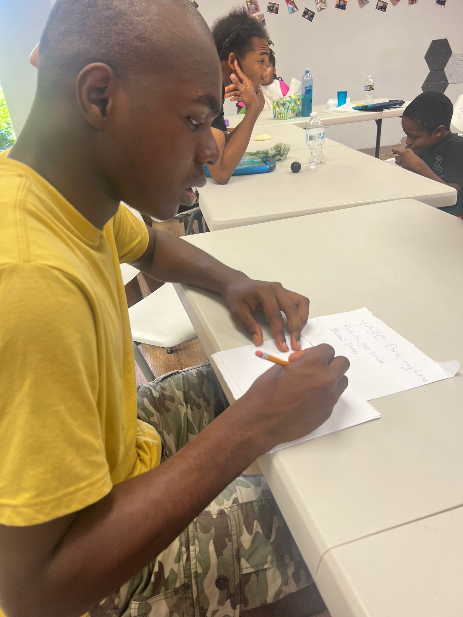 Person in yellow shirt writing at a table with others. Bright room, natural light.