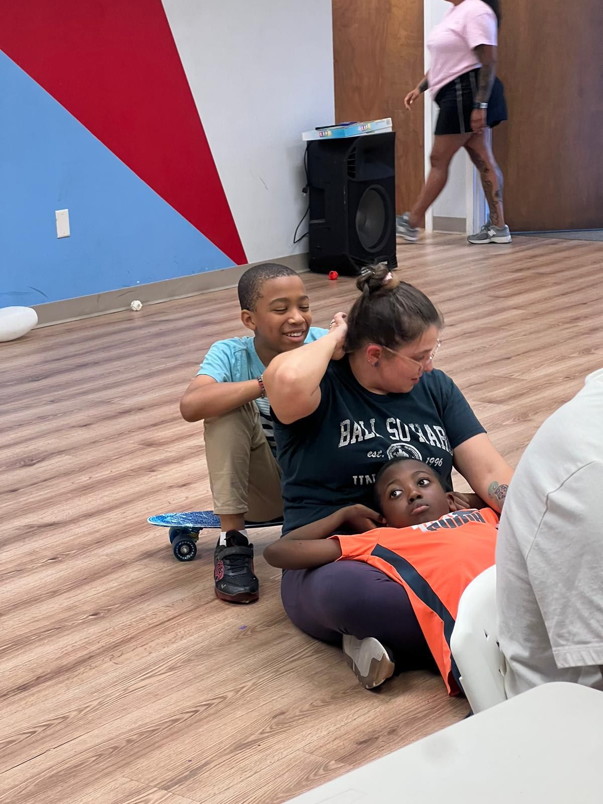 A woman and child are on the floor. A second child sits nearby on a skateboard. Someone walks in the background.