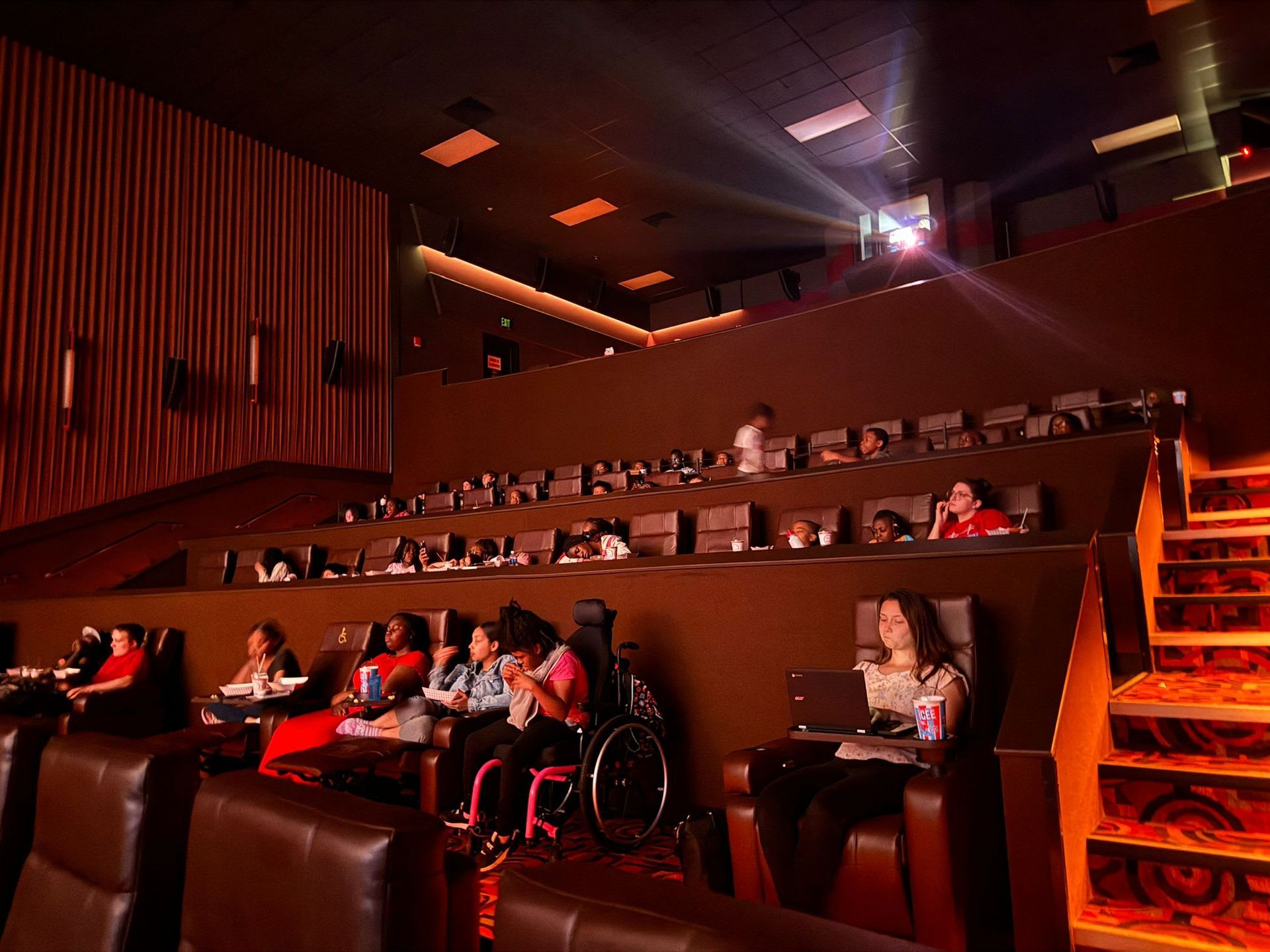 Movie theater interior with tiered seating, audience members watching, projector on ceiling.