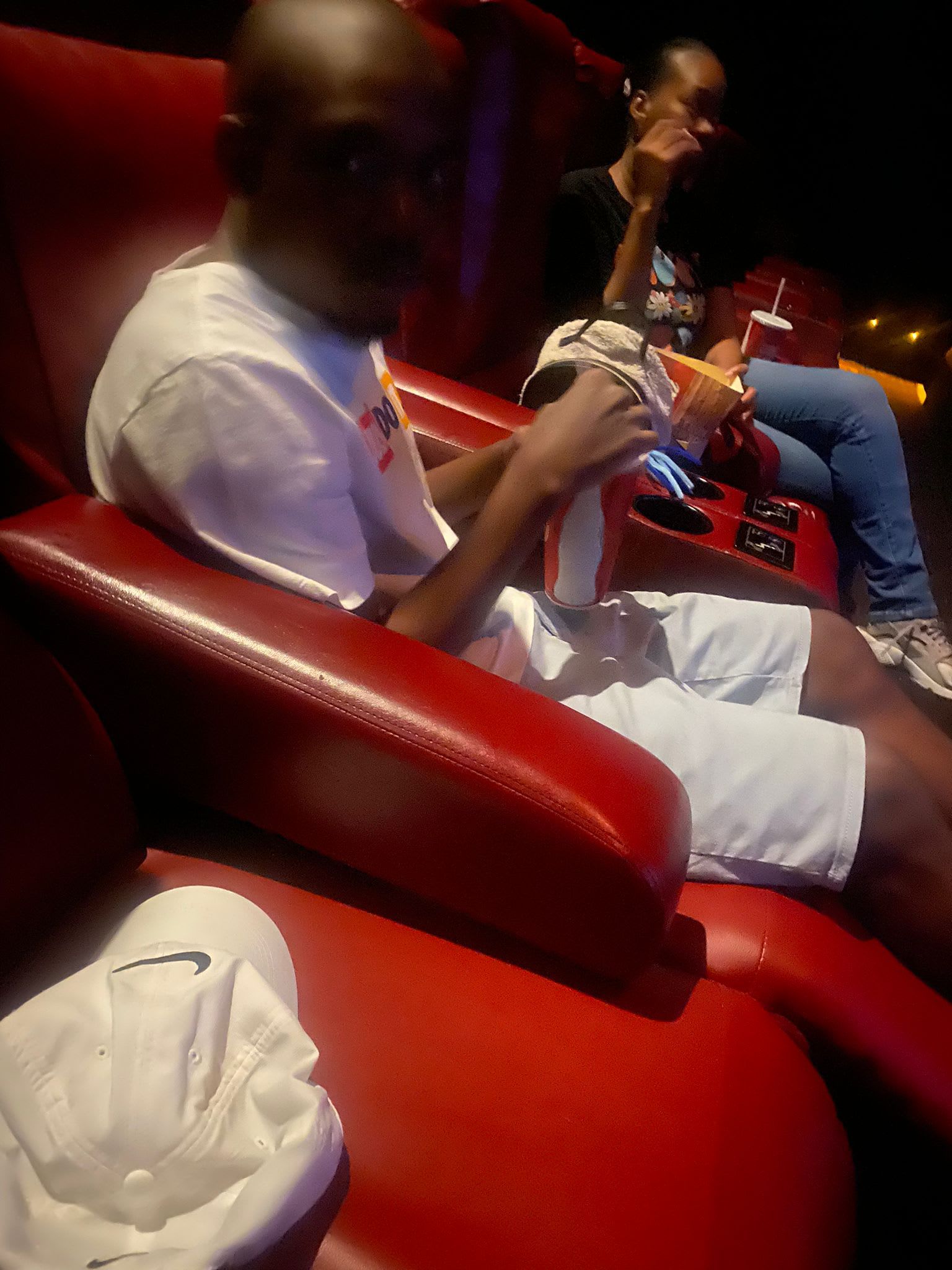 Man in white shirt and shorts sits in a red theater chair, holding food. 
