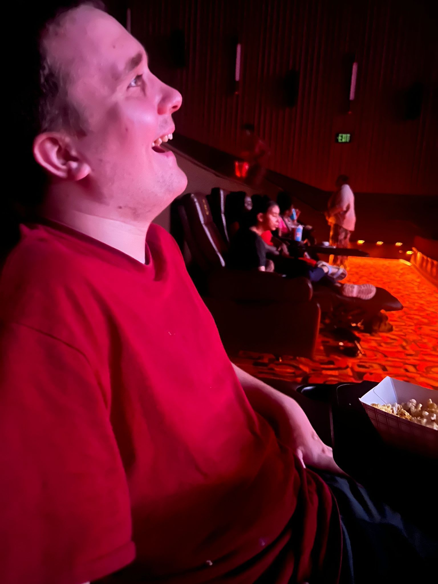 Man in red shirt laughs in a movie theater, looking upward. 