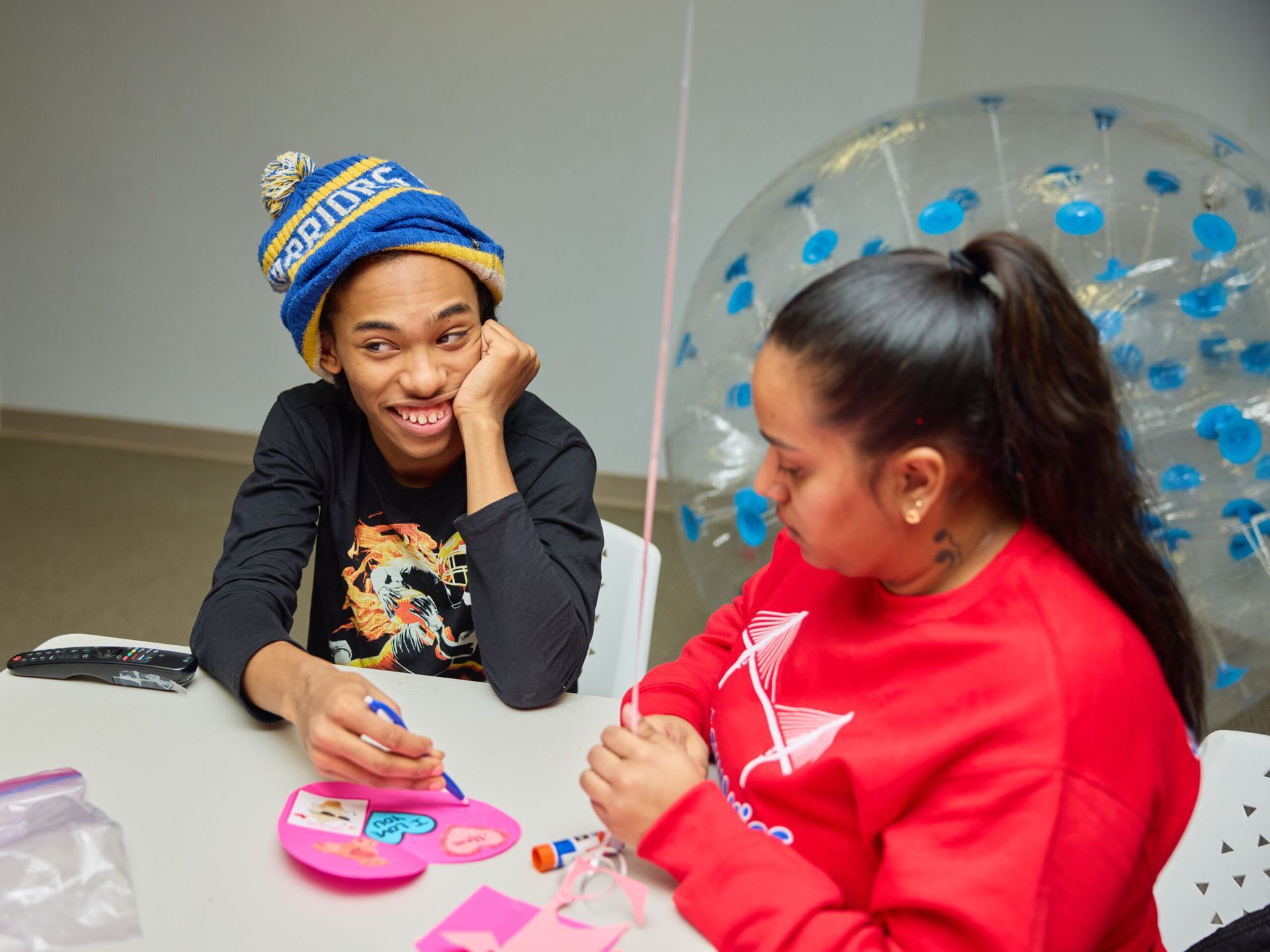 Two people crafting at a table. One smiles, wearing a blue hat. 