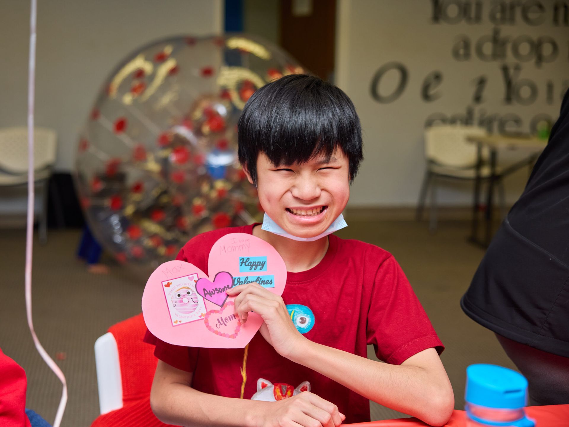 Boy holding Valentine's cards, smiling at the camera, wearing a red shirt, inside a room with a bubble.