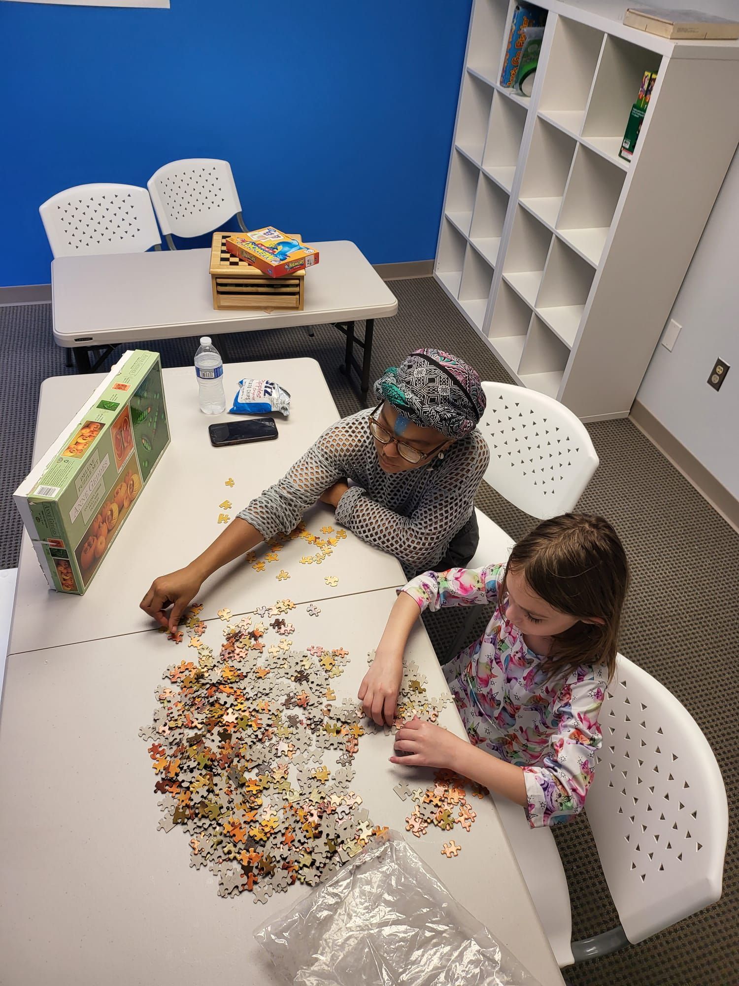 Two people working on a jigsaw puzzle at a table in a room with white shelves and blue wall.