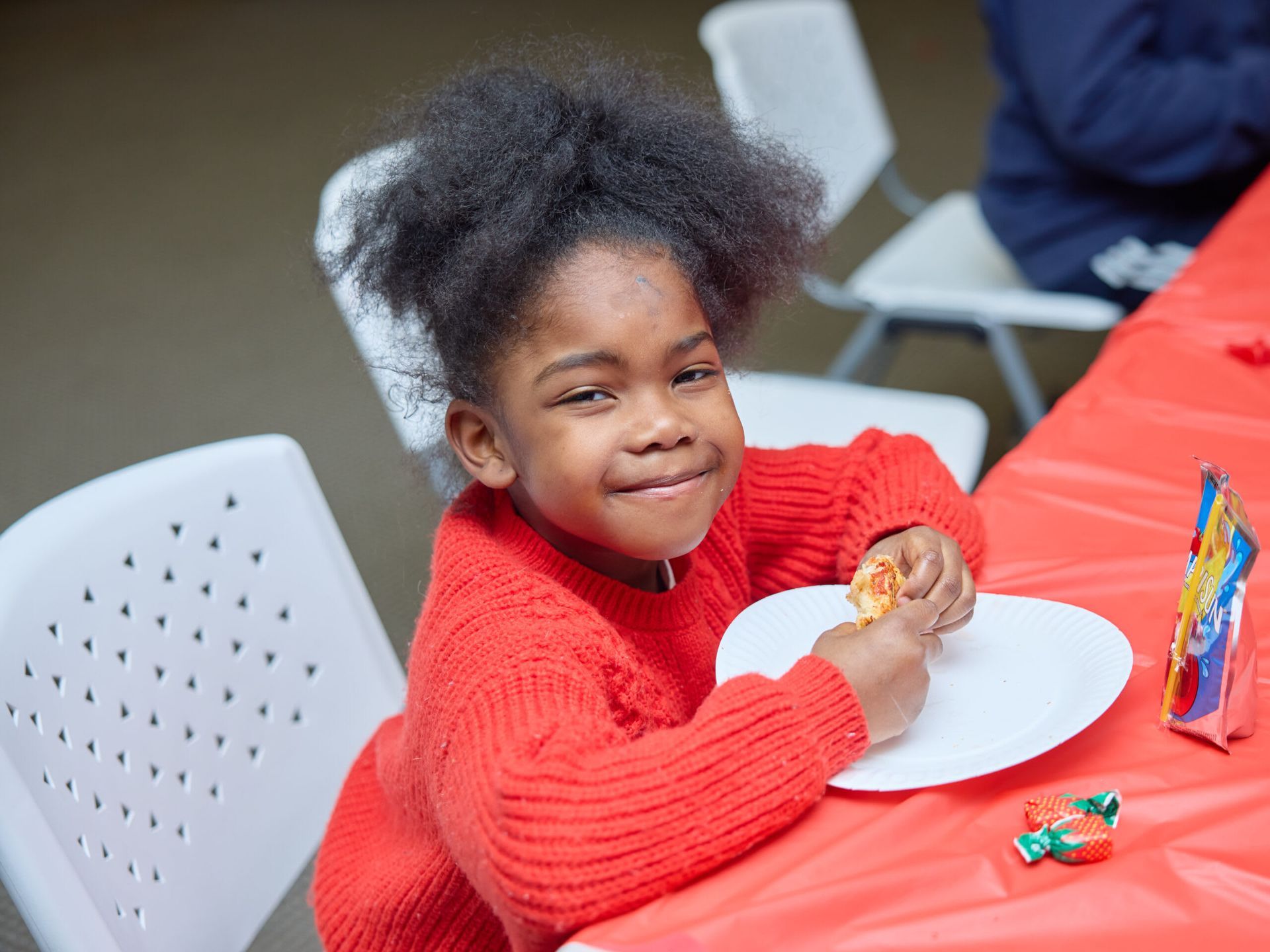 A child with curly hair smiles while eating at a red-covered table.