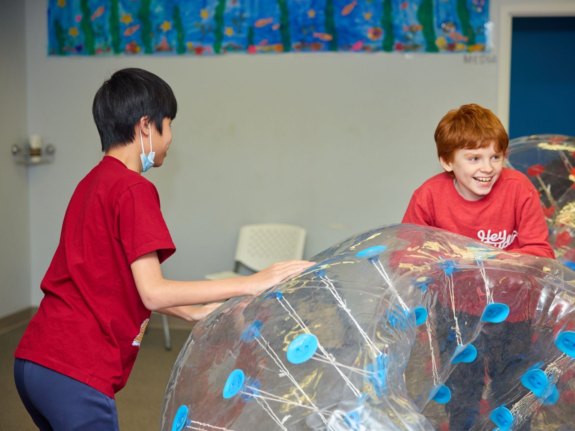 Two children playing with bubble balls in a room, one with a face mask and a red shirt.
