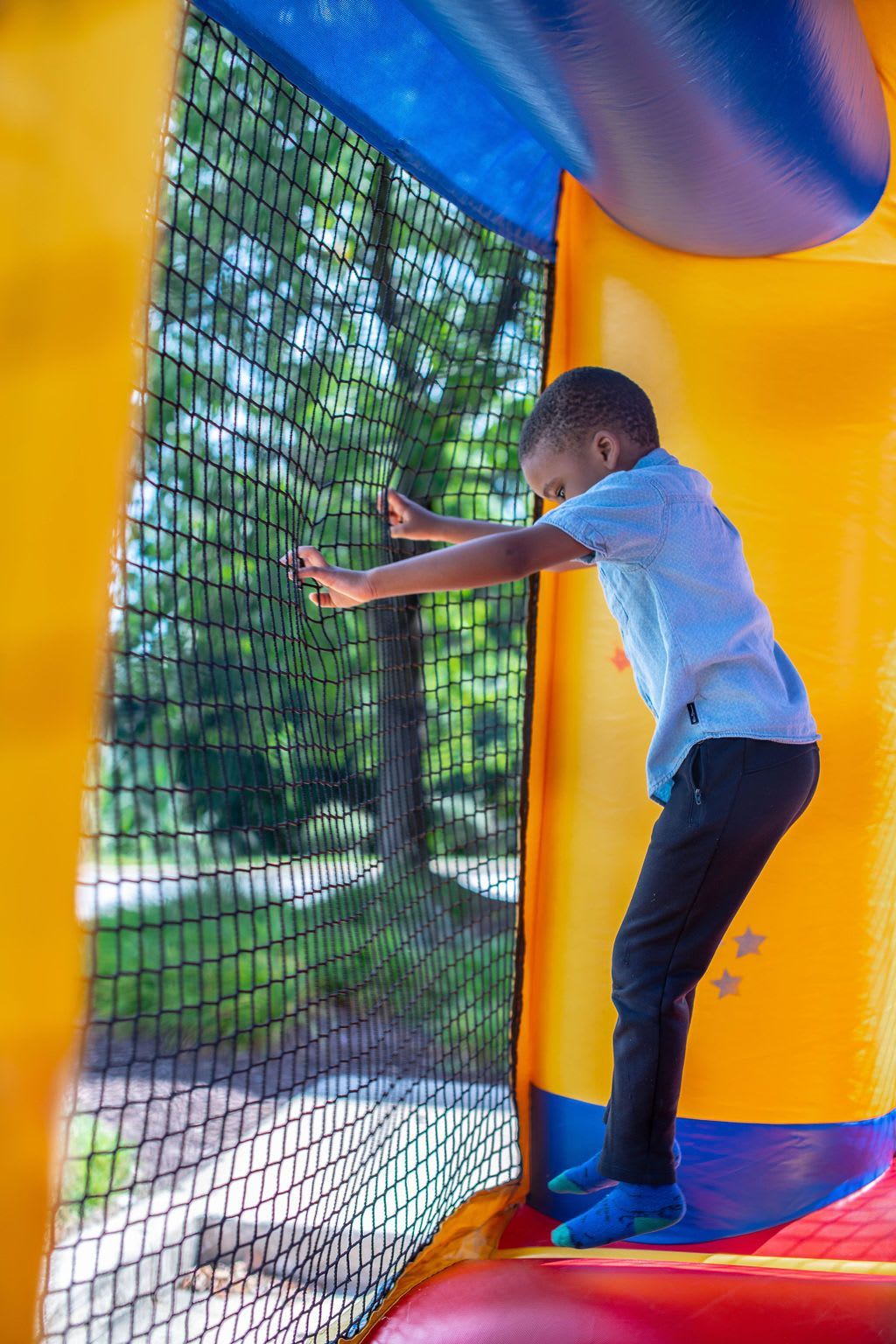 Boy in blue shirt and black pants standing inside an inflatable bounce house, looking out a mesh window.