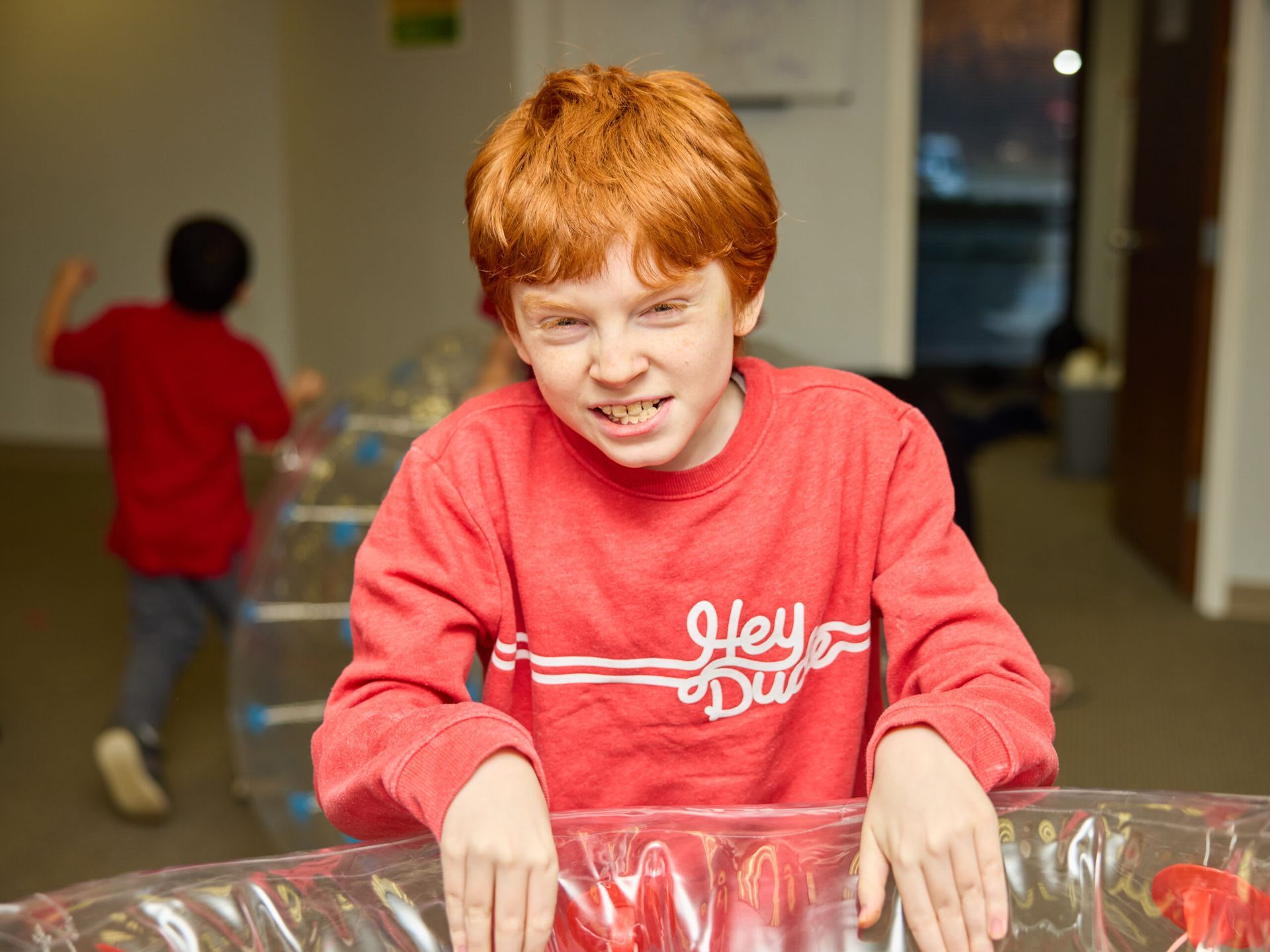 Boy with red hair in red sweatshirt smiles, leaning over clear container. 