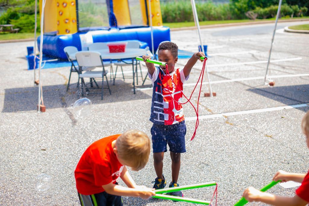 Children playing with bubble wands outdoors, near an inflatable obstacle course.