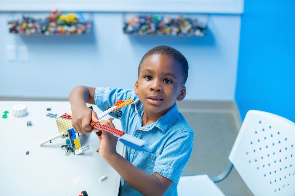Boy in blue shirt building with LEGOs at a white table in a room with blue walls, smiling at camera.