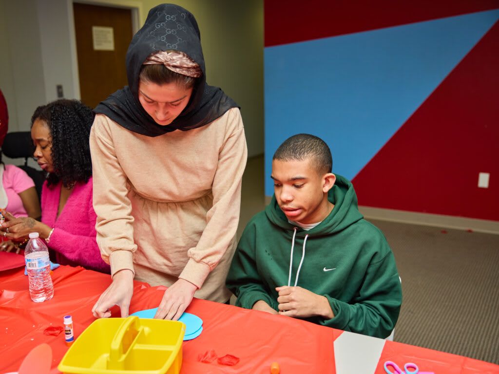 Woman in headscarf helps person craft at a table with others.