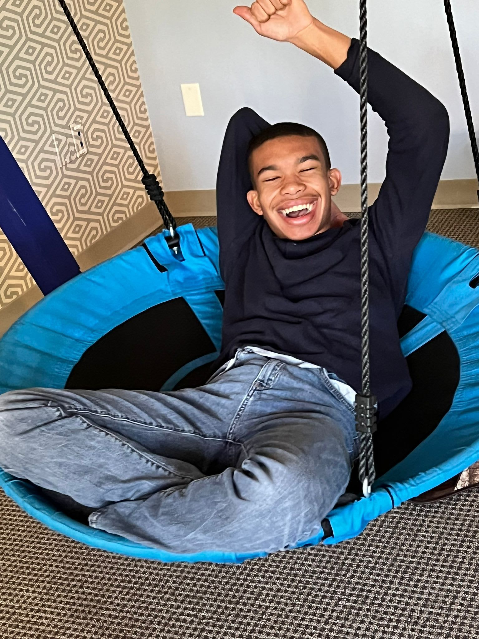 Person smiling and raising arm in a blue swing, wearing a dark blue shirt and jeans, indoors.