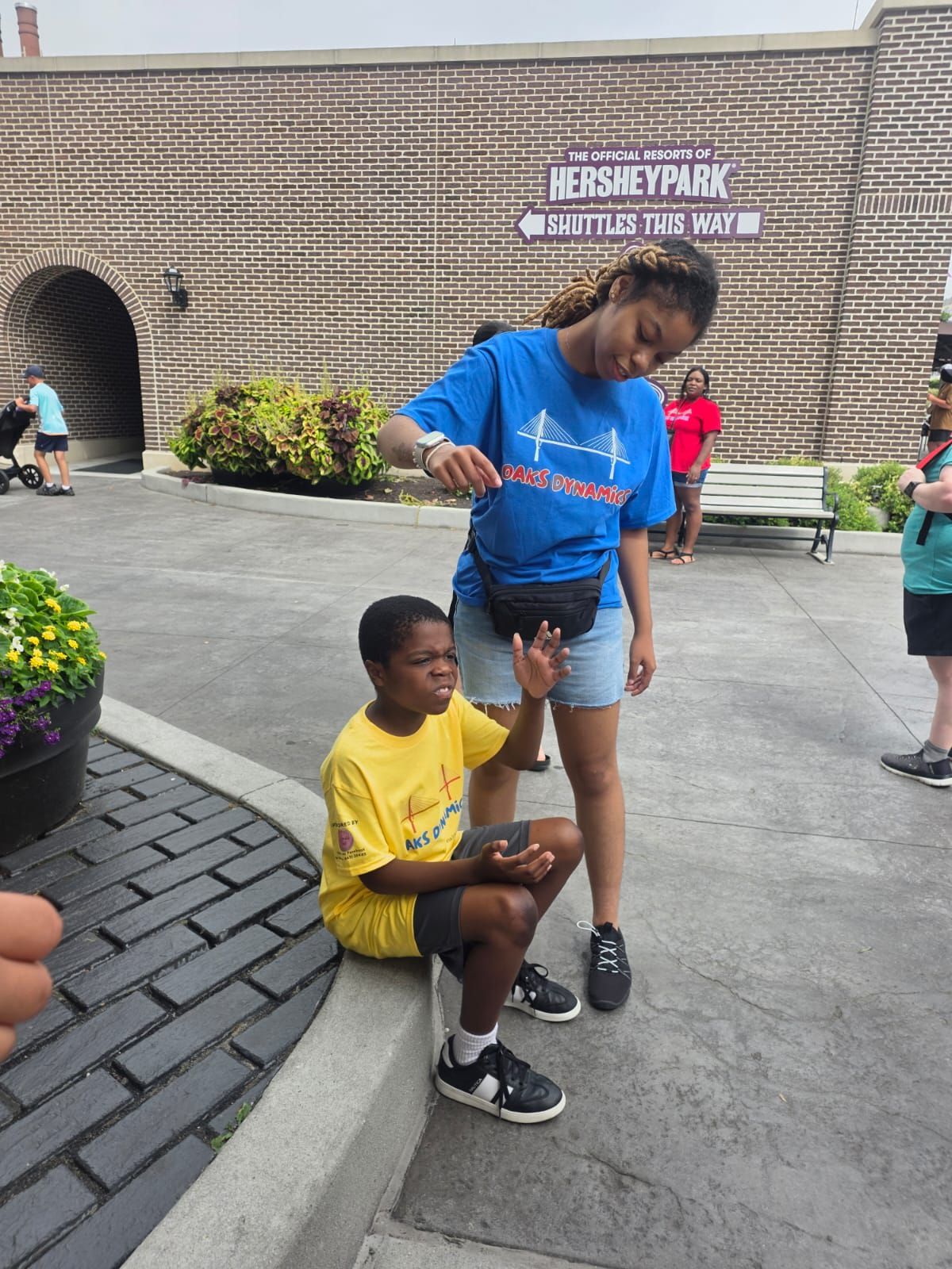 A young person in yellow shirt sits, while another in blue shirt gestures near Hershey Park sign.