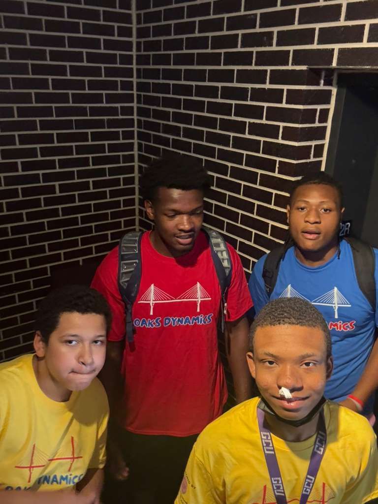 Four people wearing colorful shirts stand near a brick wall.
