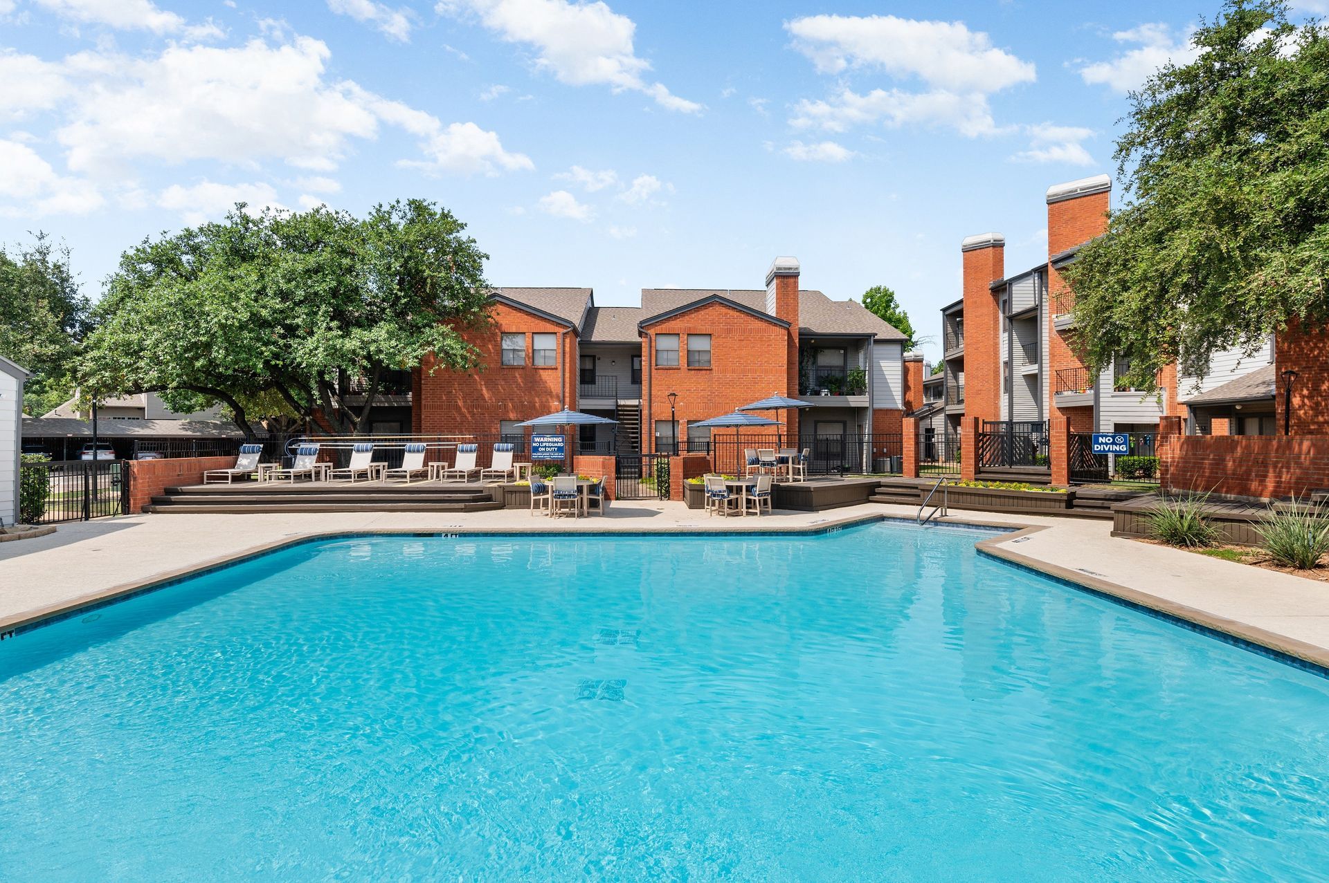 Apartment pool with clear blue water, brick buildings, lounge chairs, and trees under a blue sky.
