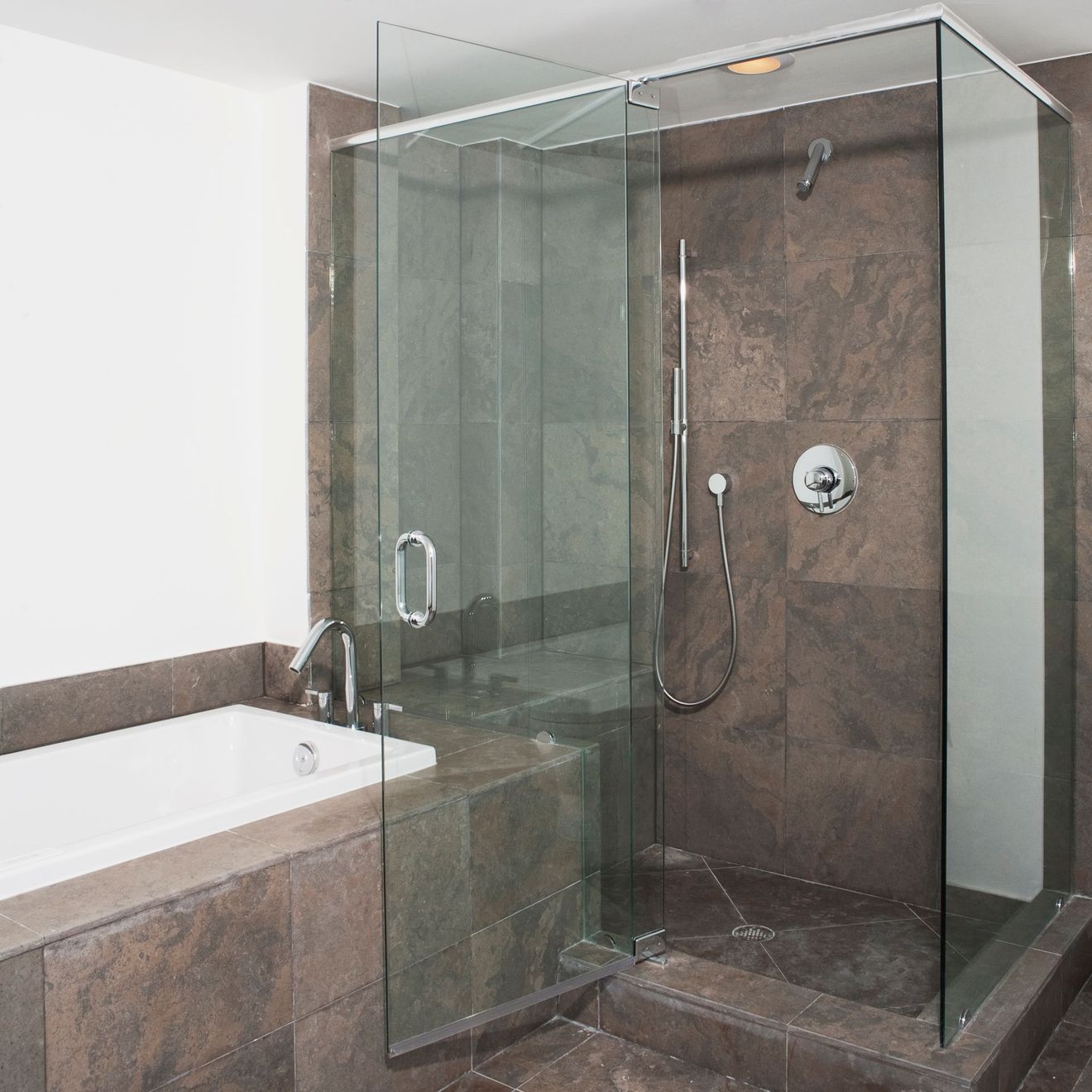 Modern bathroom with a glass shower and bathtub. Brown tile walls, white tub, clear shower door.