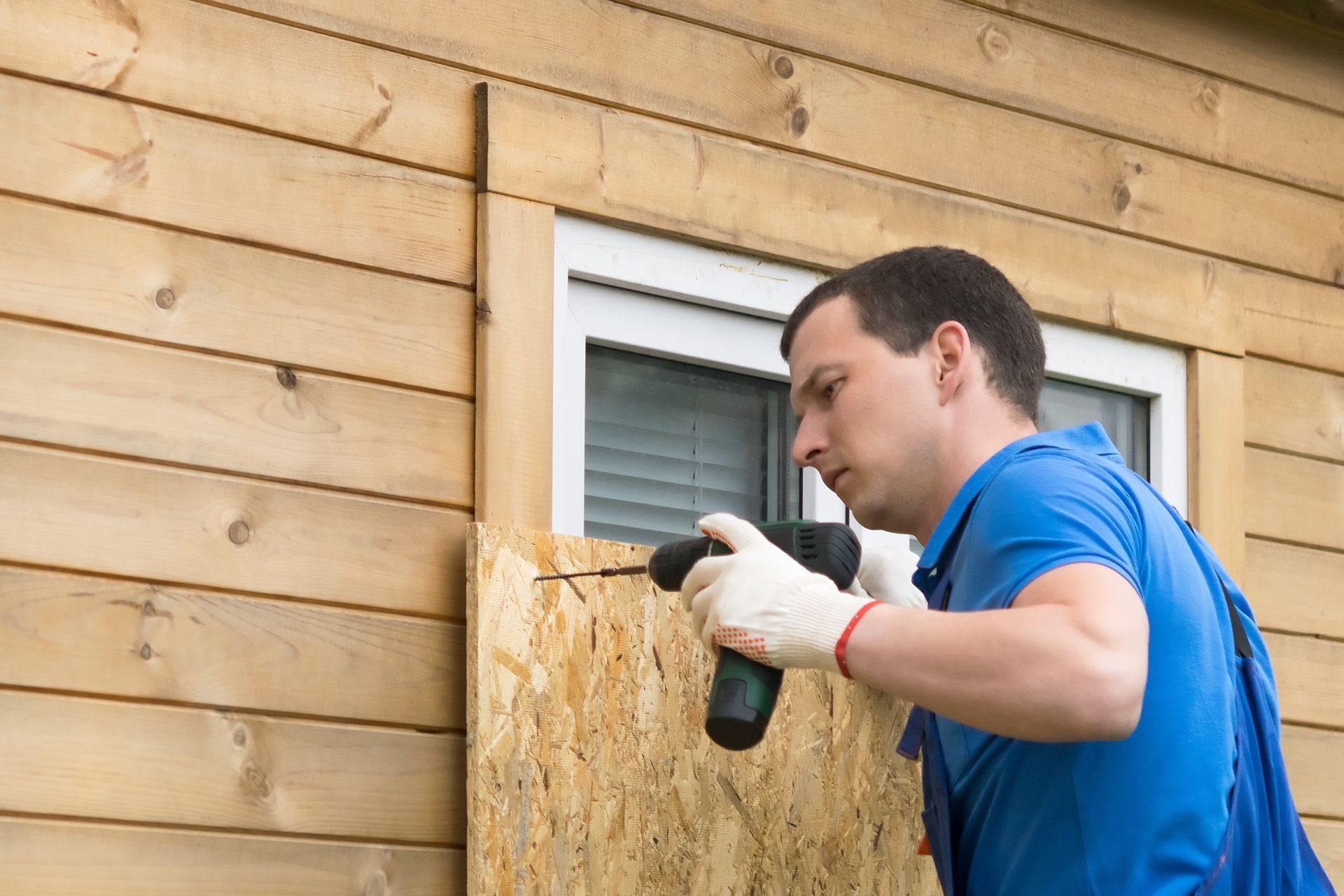 Man boards up a window with a power drill on a wooden building.