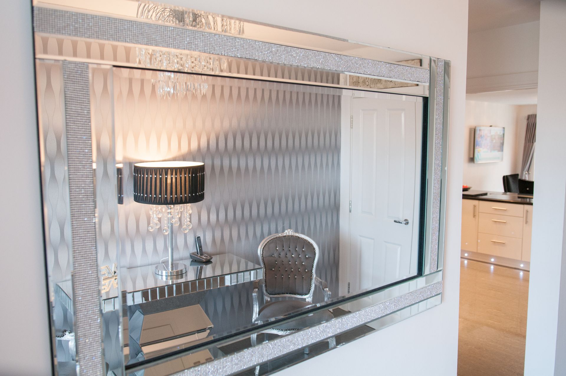 A large, ornate mirror reflecting a desk, chair, and lamp in a room with silver patterned wallpaper.
