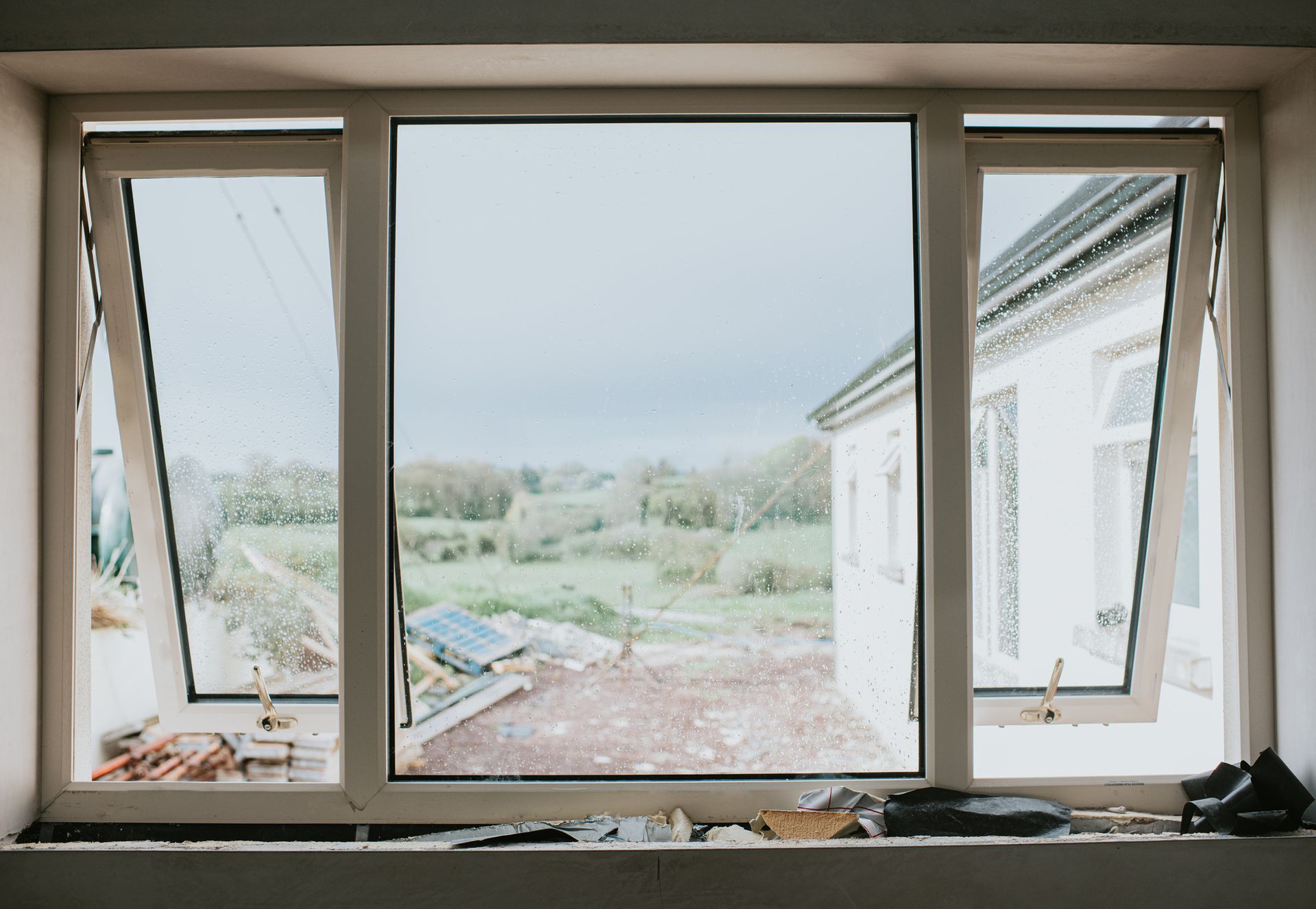 Window with open side panels overlooking a backyard with a white house and cloudy sky.
