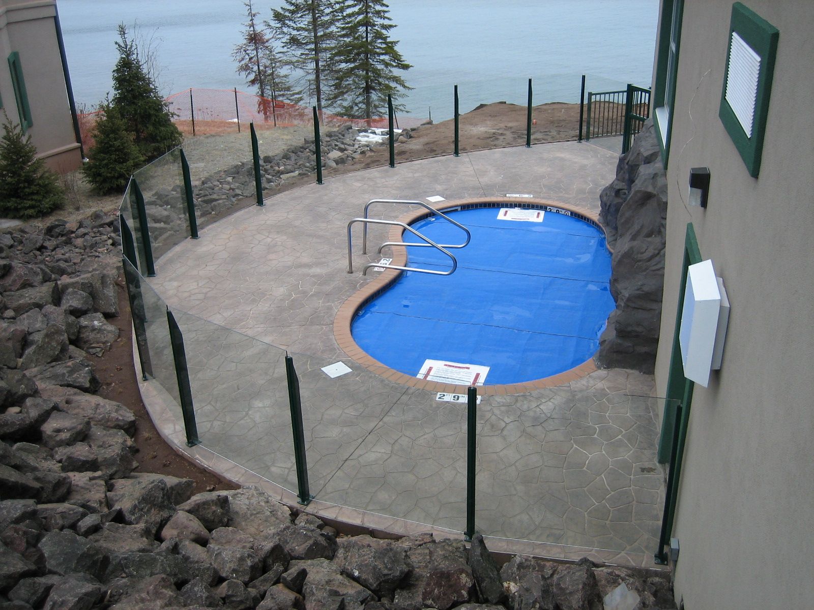 Pool with blue cover, surrounded by glass fence, concrete, and rocks. Near water and buildings.