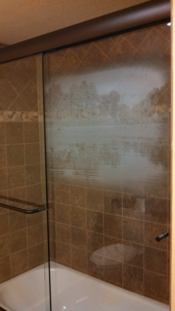 Shower with a glass door. Brown tile walls and a white tub. Scenic frosted design on the glass.