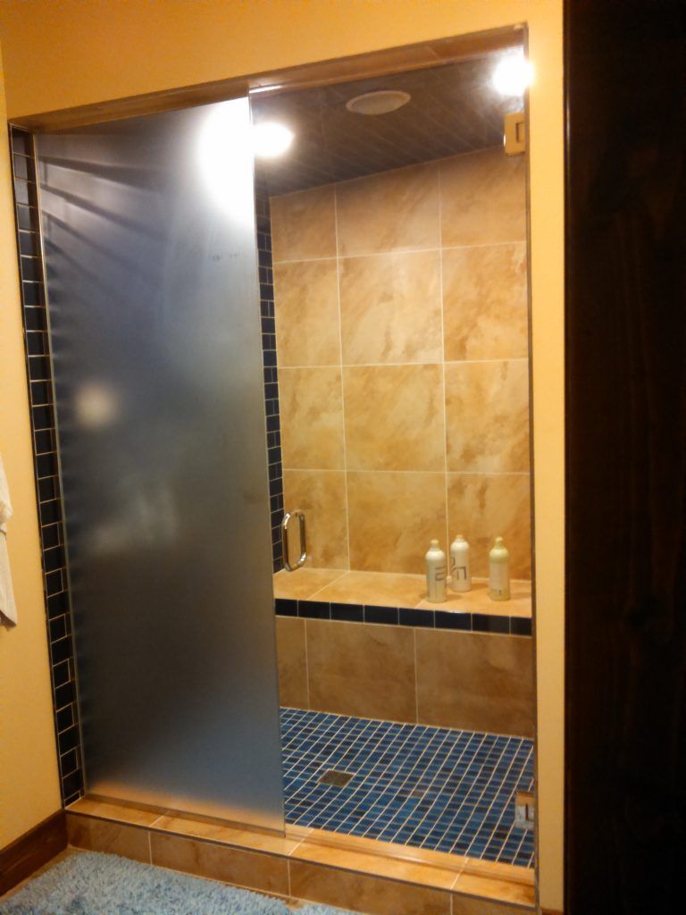 Shower with frosted glass door, tan tile walls, blue mosaic floor, and a bench.
