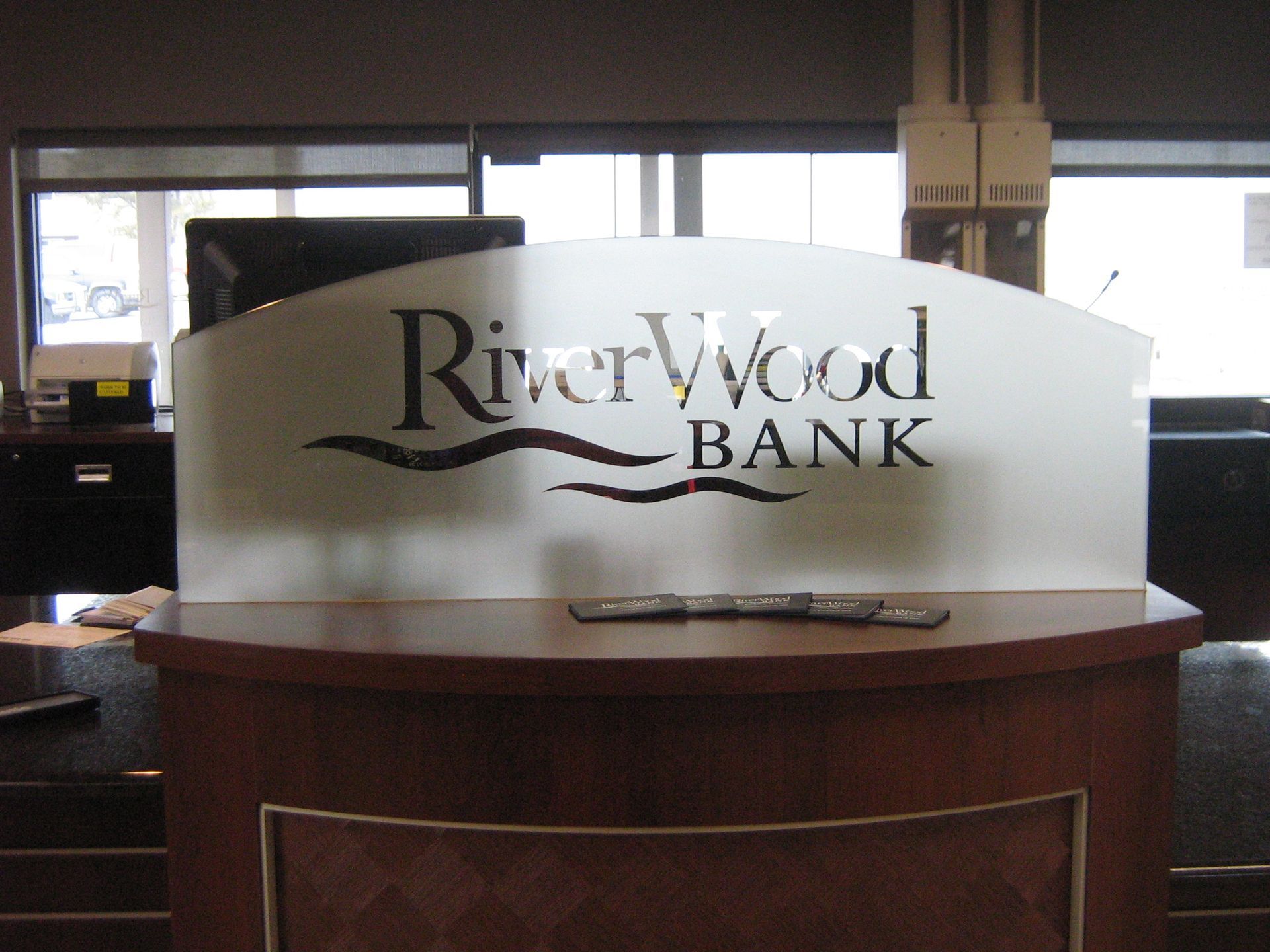RiverWood Bank reception desk with white logo on a curved, frosted glass shield.