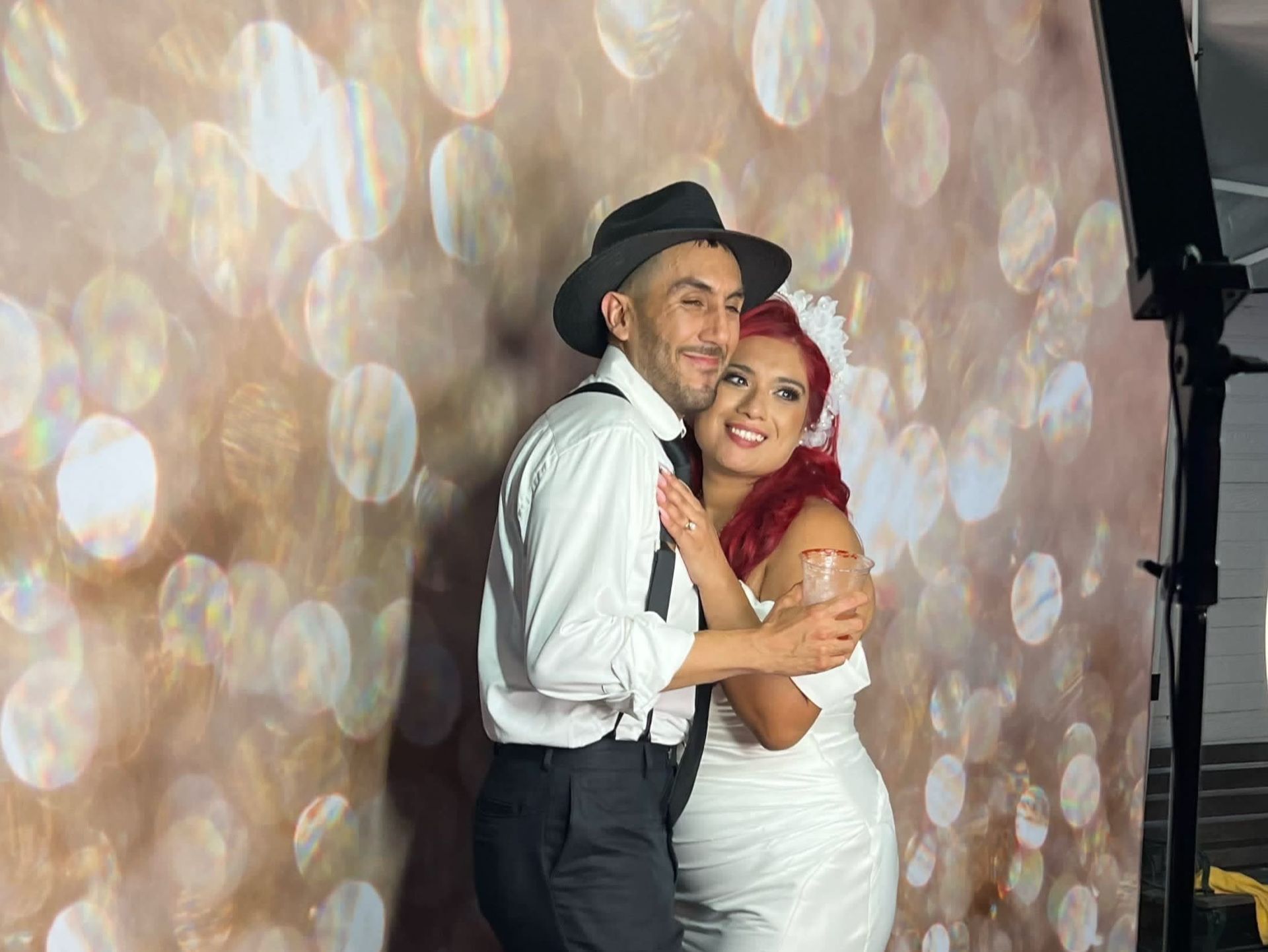 Couple embracing in front of a bokeh backdrop in a photo booth; man in hat and suspenders, woman in white dress, both smiling.