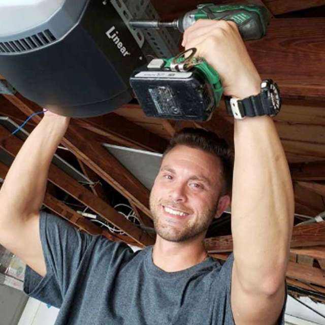 Man smiling while installing a garage door opener with a power drill in a garage.