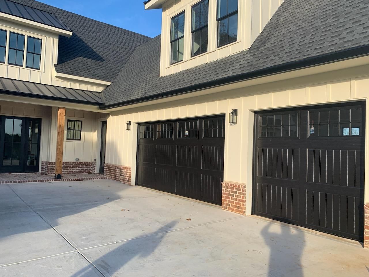Black garage doors on a light-colored house with a brick base, a concrete driveway, and a blue sky.