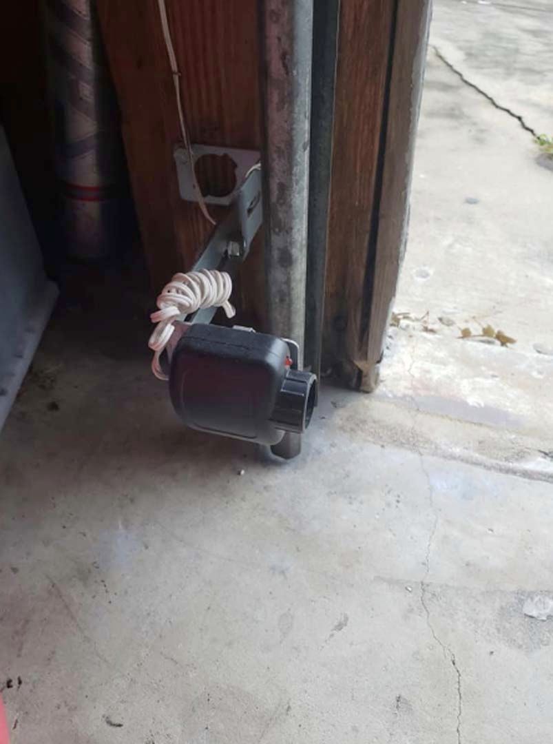 Garage door sensor, black, mounted on a metal bracket, with white cord. Concrete floor.