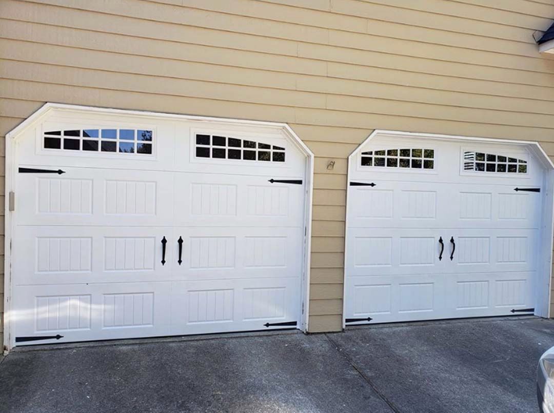 Two white garage doors with decorative windows and black handles, on a tan house.