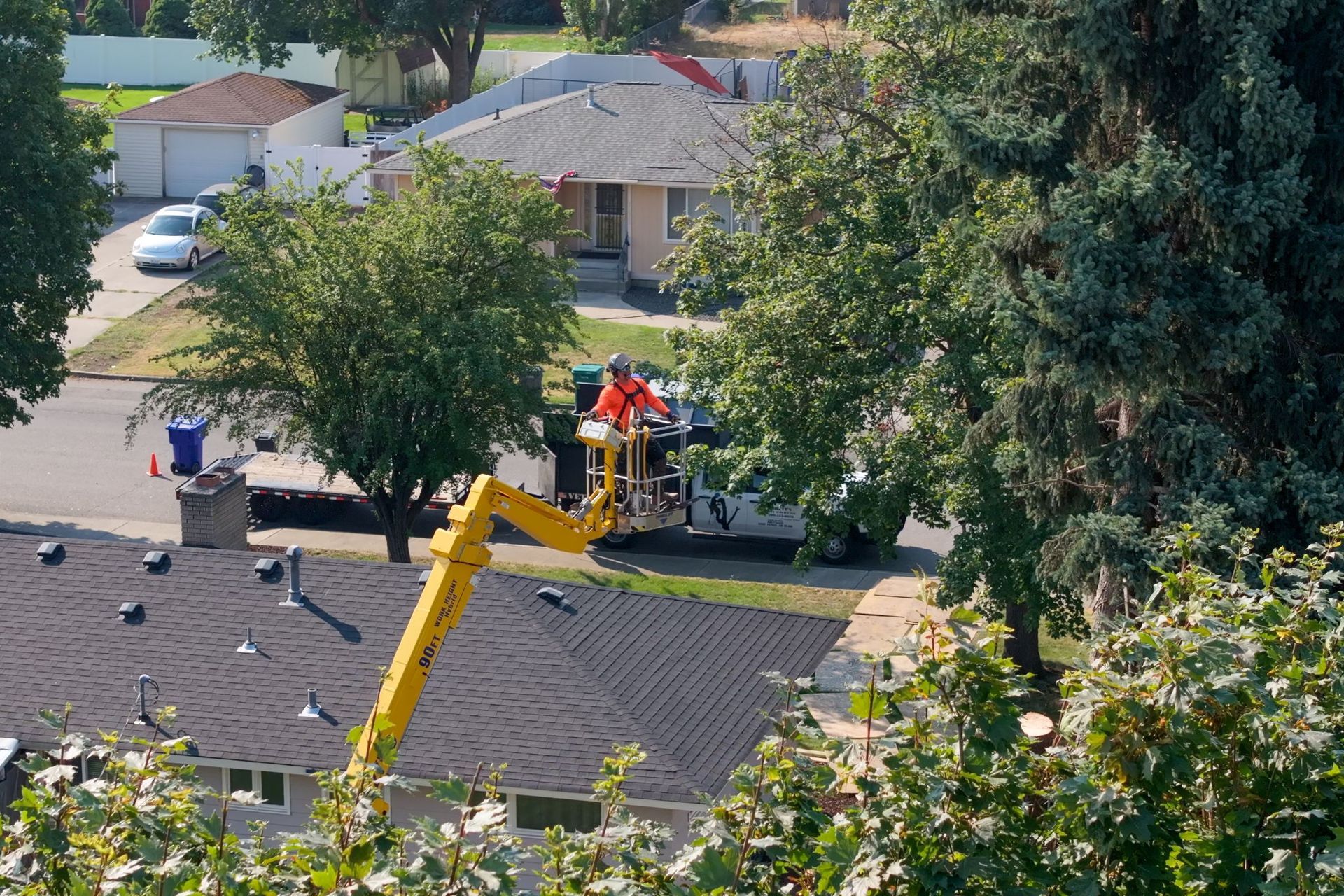 Yellow lift truck with worker trimming a tree near houses.