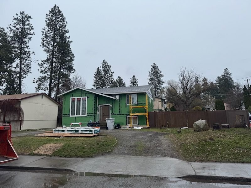 A residential house under exterior renovation with exposed green sheathing, construction materials, and a fenced yard.