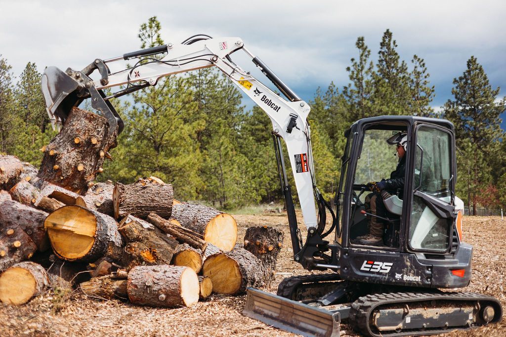 Mini excavator loading logs. A person operates the machine in an outdoor setting.