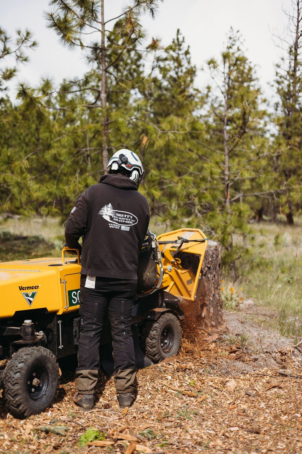Person operating a yellow stump grinder in a wooded area, wearing protective gear.