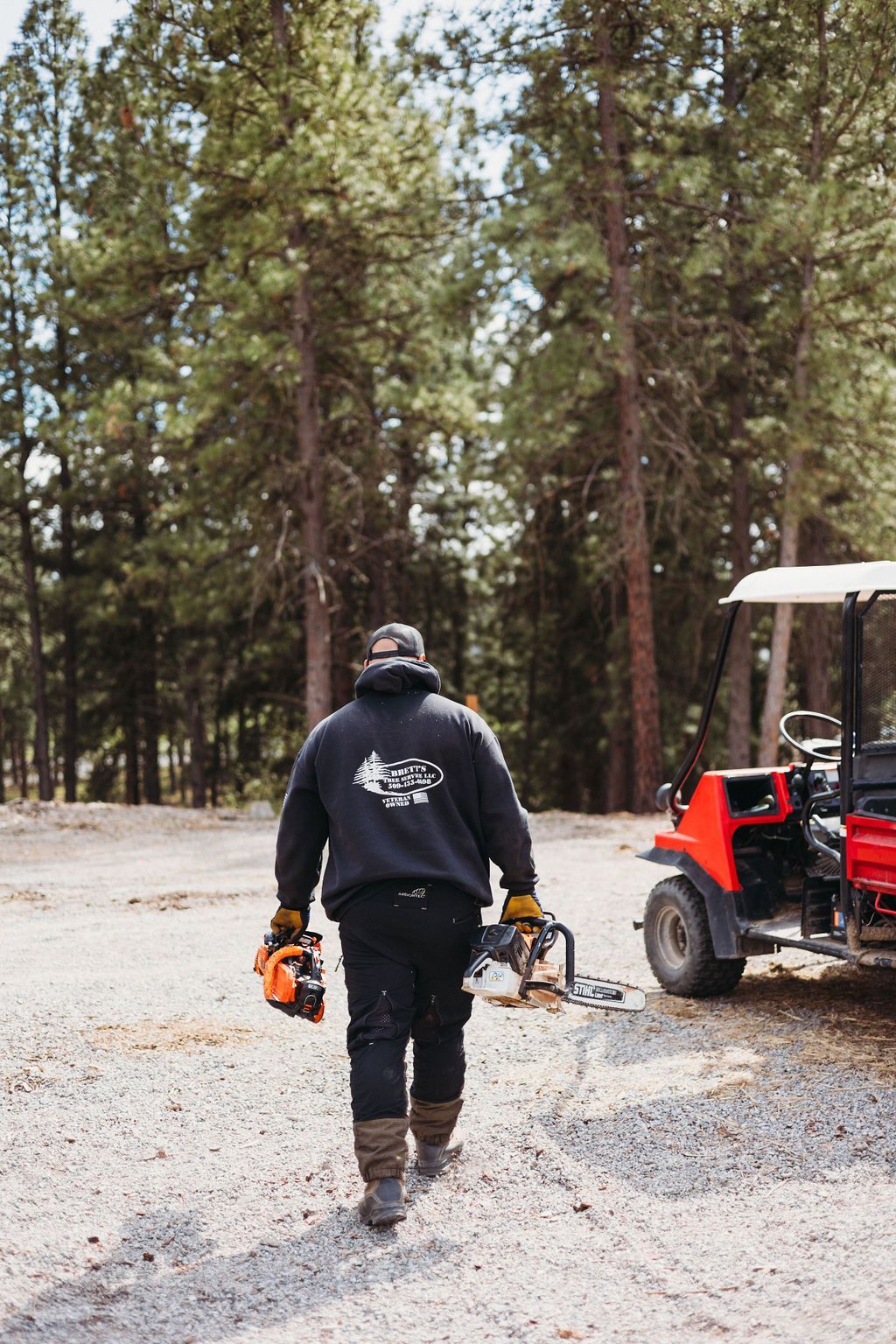 Man walking away, carrying two chainsaws, toward trees and red utility vehicle on gravel path.