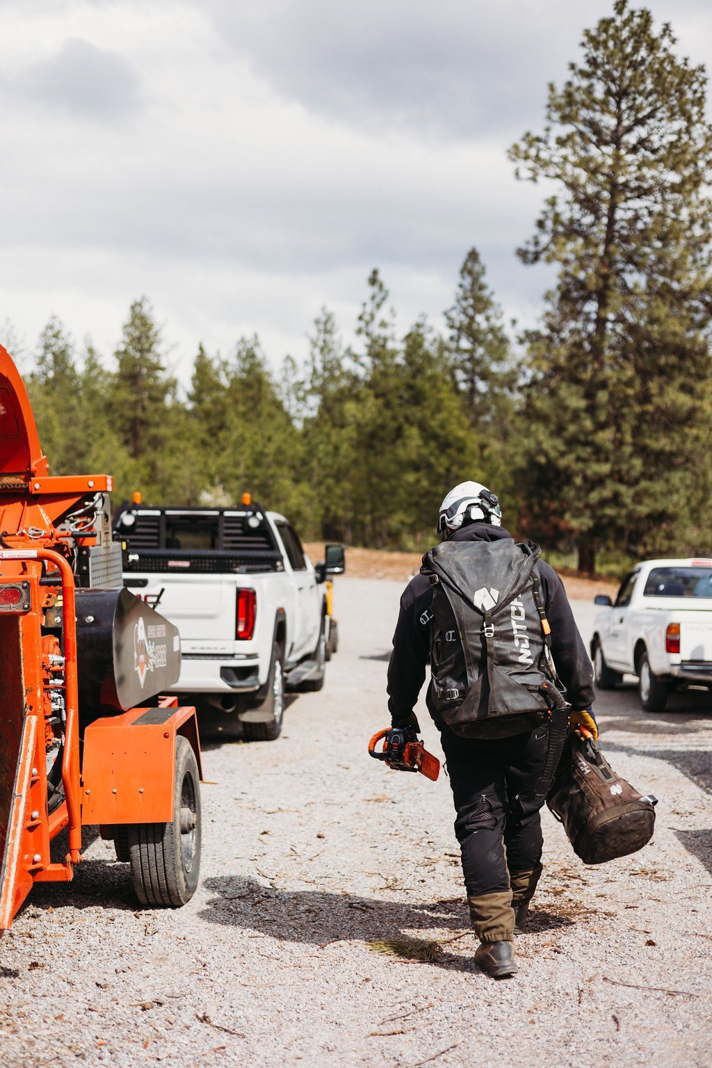 A person in work gear walks toward a wood chipper and trucks in a gravel area; pine trees in background.