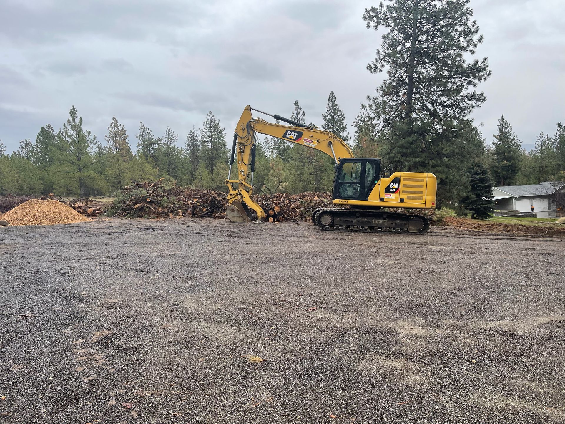 Yellow excavator at a gravel-covered site; a large pile of wood chips and trees in the background. Overcast sky.