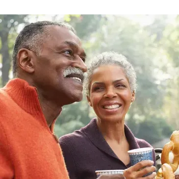 Smiling couple enjoying an outdoor event; man in orange shirt, woman with coffee cup.