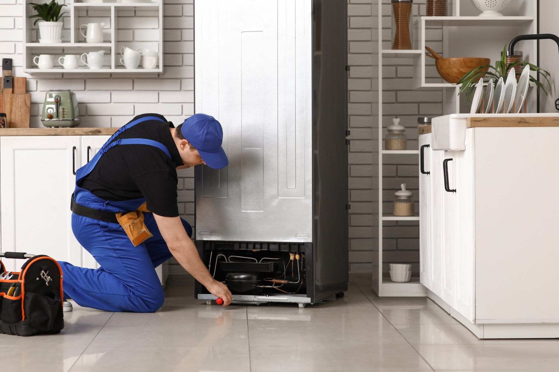 A repairman in blue overalls kneels to fix a refrigerator in a kitchen with white cabinets.